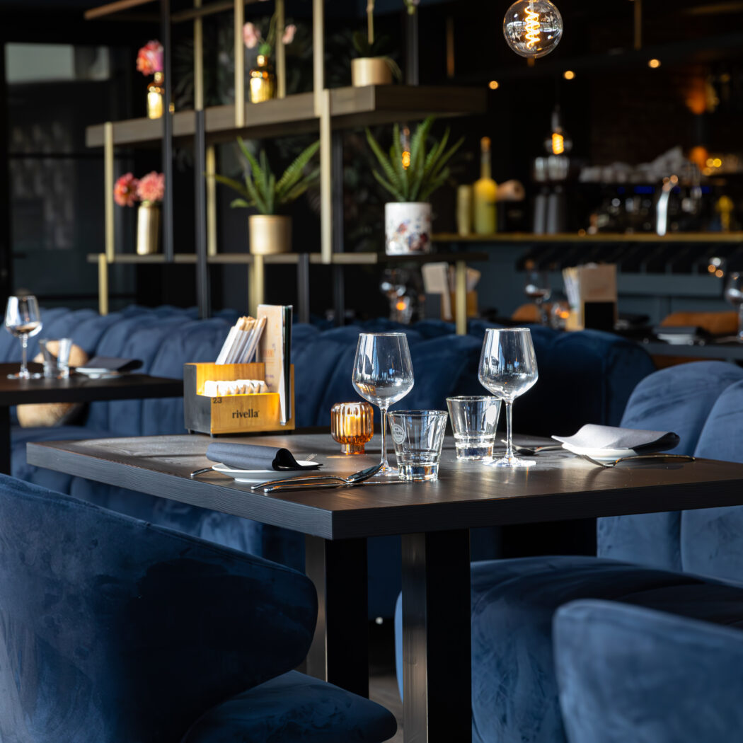 Sfeervol restaurant met blauwe fluwelen banken en stoelen, gedekte tafels, en een Rivella menuhouder.