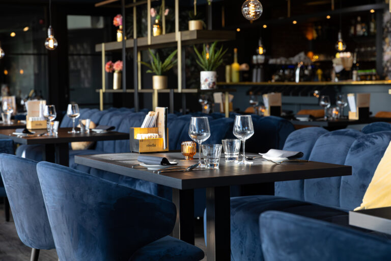 Sfeervol restaurant met blauwe fluwelen banken en stoelen, gedekte tafels, en een Rivella menuhouder.