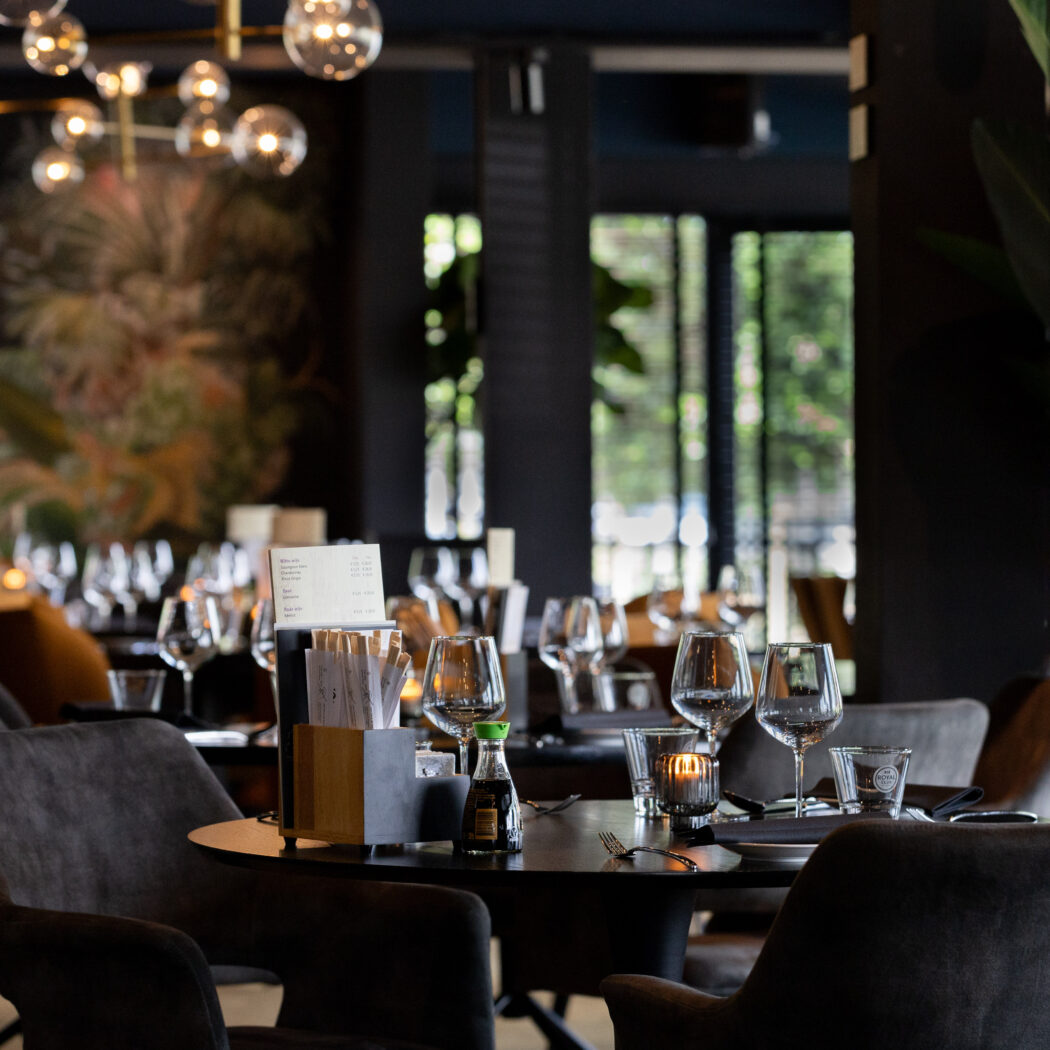Donker, sfeervol restaurantinterieur met gedekte tafels, glazen, kaarsen en een kleurrijk schilderij.