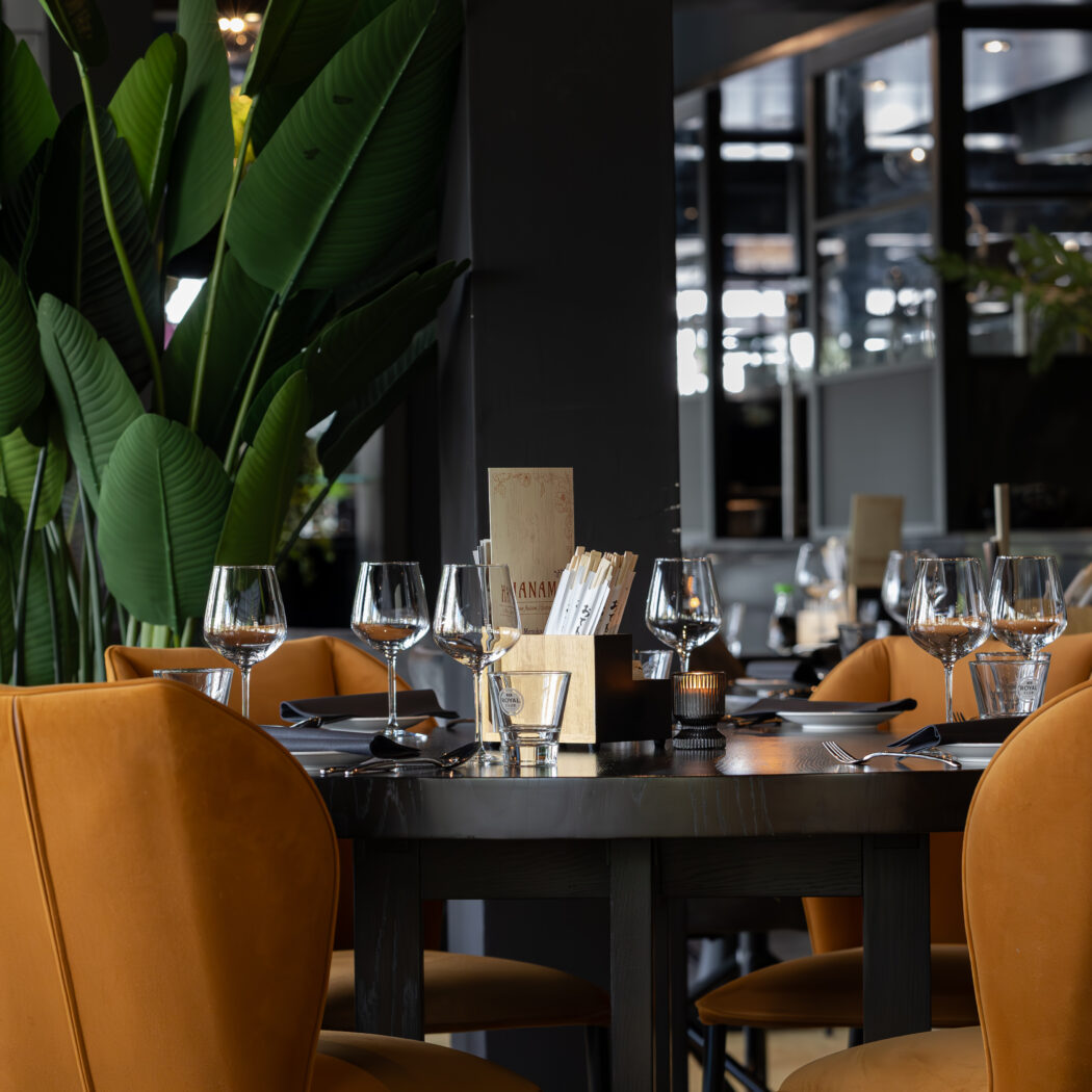 Gedekte restauranttafel met okergele velours stoelen, wijnglazen en menu met HANAM, omringd door grote groene planten.