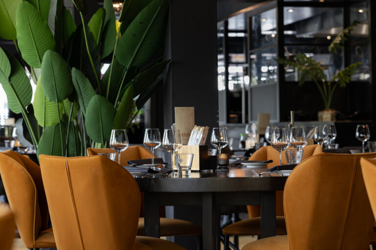 Gedekte restauranttafel met okergele velours stoelen, wijnglazen en menu met HANAM, omringd door grote groene planten.