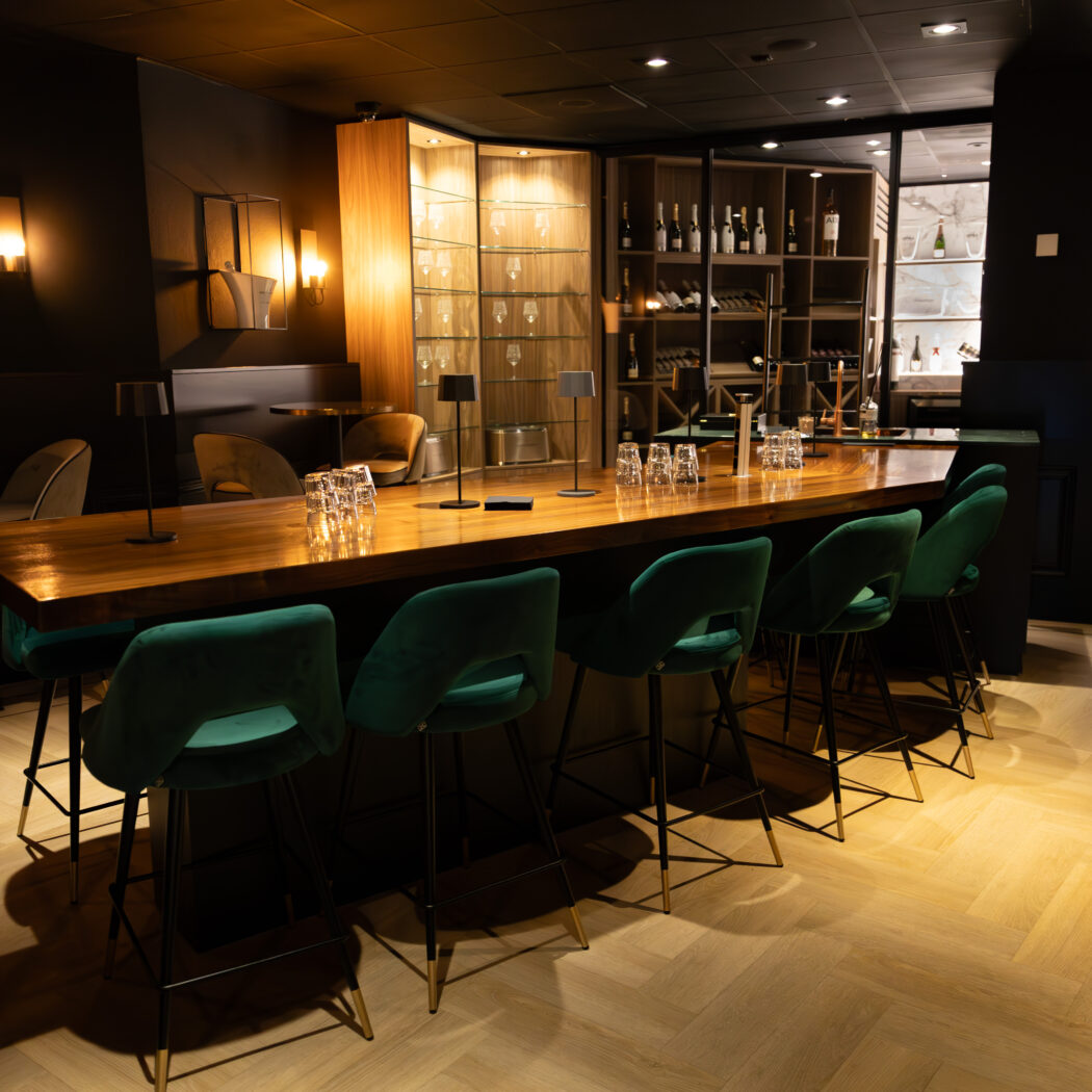 Stijlvolle, donkere bar met lange houten tafel, groene fluwelen barkrukken en verlichte schappen.