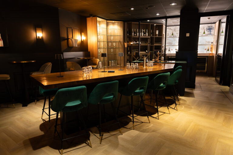 Stijlvolle, donkere bar met lange houten tafel, groene fluwelen barkrukken en verlichte schappen.