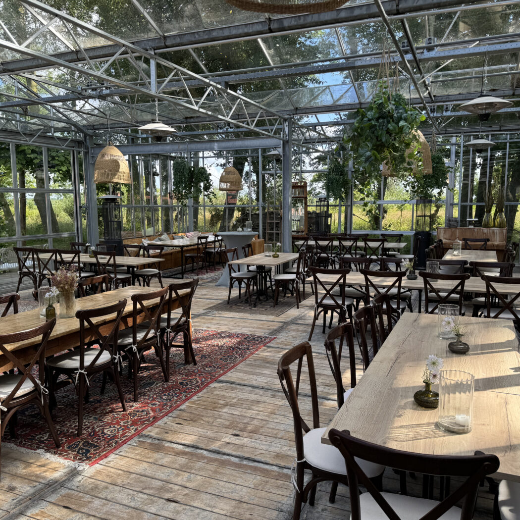 Gezellige evenementenruimte in glazen kas met houten tafels en kruisrugstoelen, omringd door natuur.