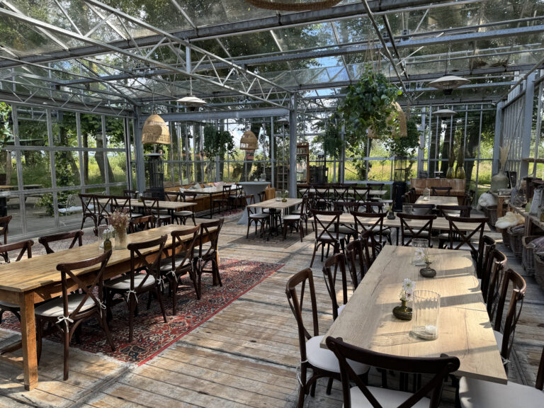 Gezellige evenementenruimte in glazen kas met houten tafels en kruisrugstoelen, omringd door natuur.