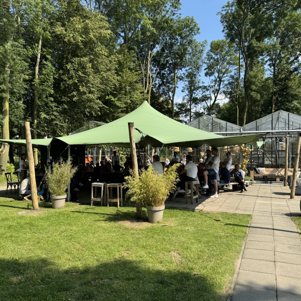 Groene stretchtent vol mensen in een weelderige buitenomgeving met glazen kassen.