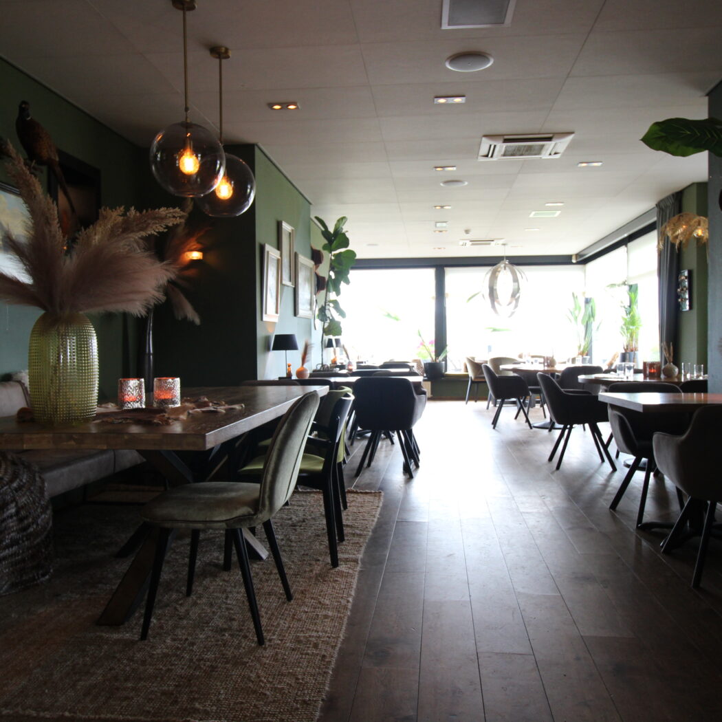 Gezellige eetruimte met donkergroene muren, houten tafels, velvet stoelen, pampasgras en glazen hanglampen.