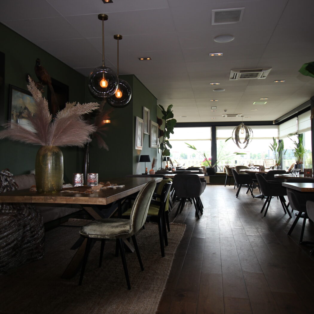 Sfeervol restaurantinterieur met donkergroene muren, houten vloer, een lange tafel met pampasgras en moderne hanglampen.