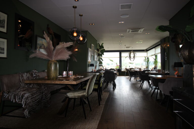 Sfeervol restaurantinterieur met donkergroene muren, houten vloer, een lange tafel met pampasgras en moderne hanglampen.