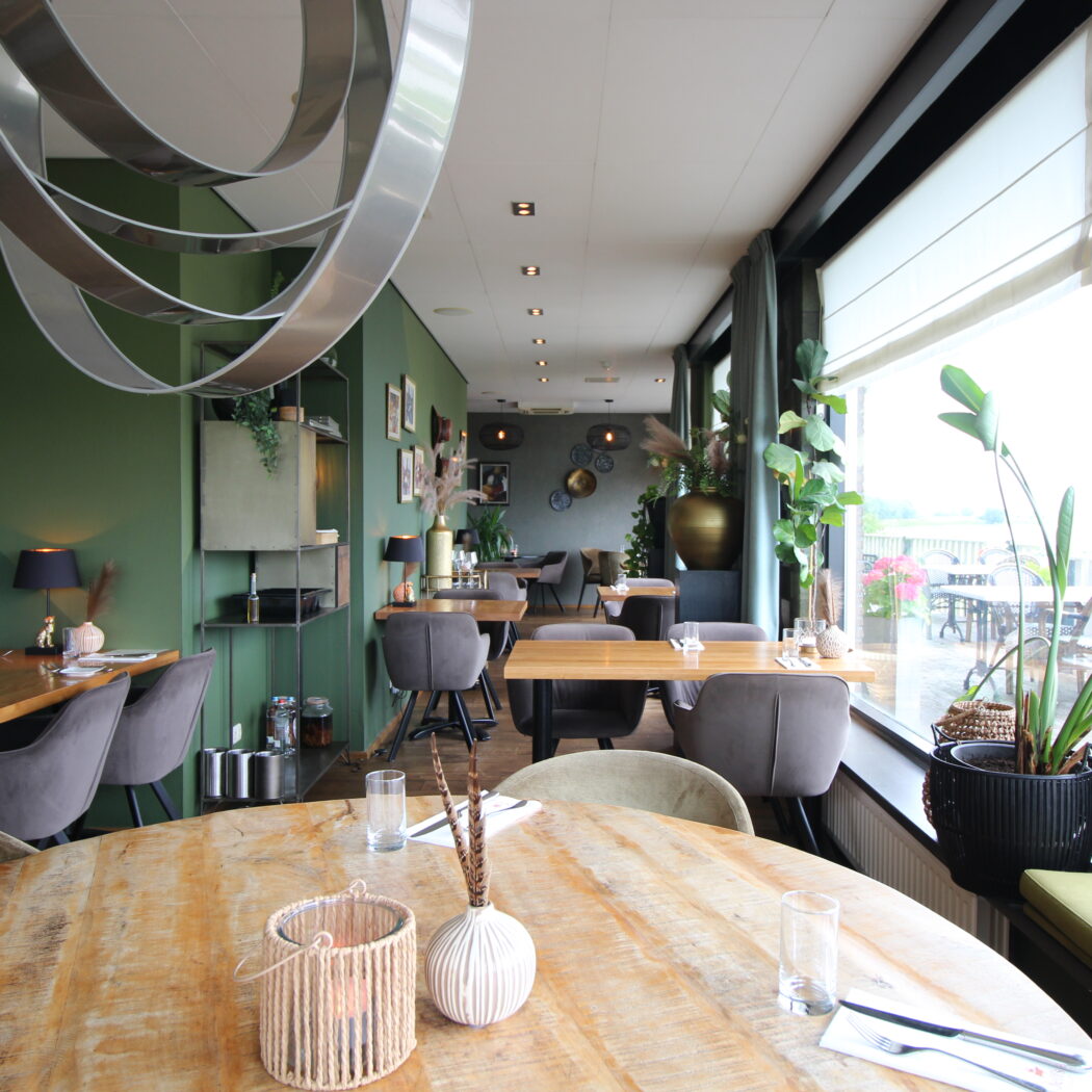 Sfeervol restaurant interieur met groene muren, houten tafels en grote ramen die uitkijken op een buitenterras.