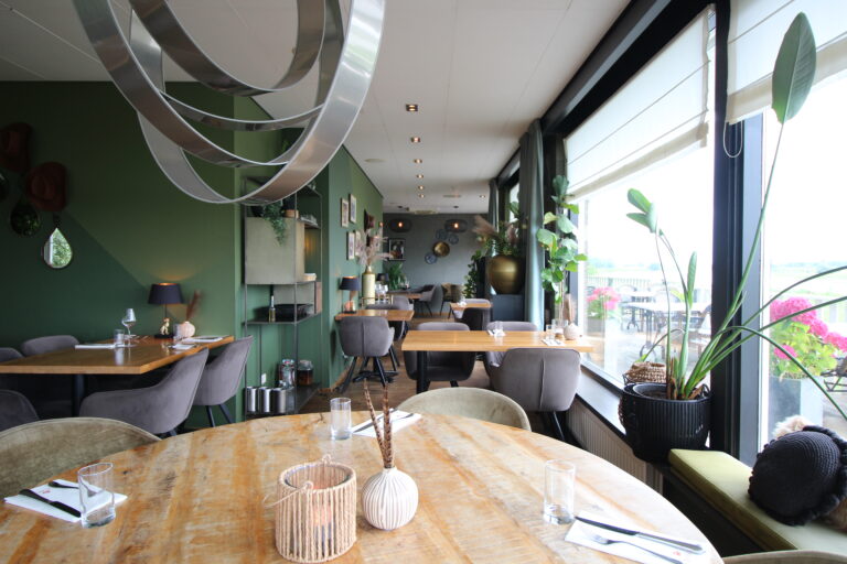 Sfeervol restaurant interieur met groene muren, houten tafels en grote ramen die uitkijken op een buitenterras.