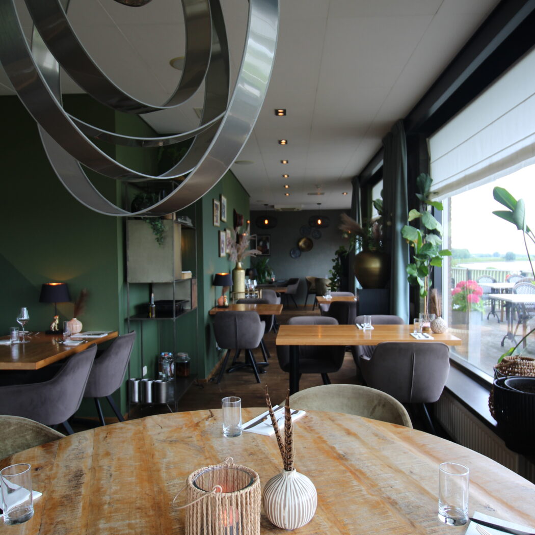 Sfeervol restaurant met houten tafels, groene muren, een grote metalen hanglamp en uitzicht op groen landschap.