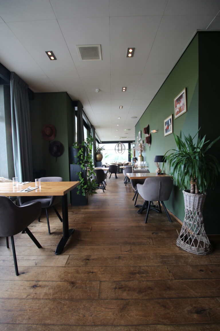 Lange restaurantruimte met donkergroene muren, houten vloer, moderne tafels, grijze stoelen en planten.