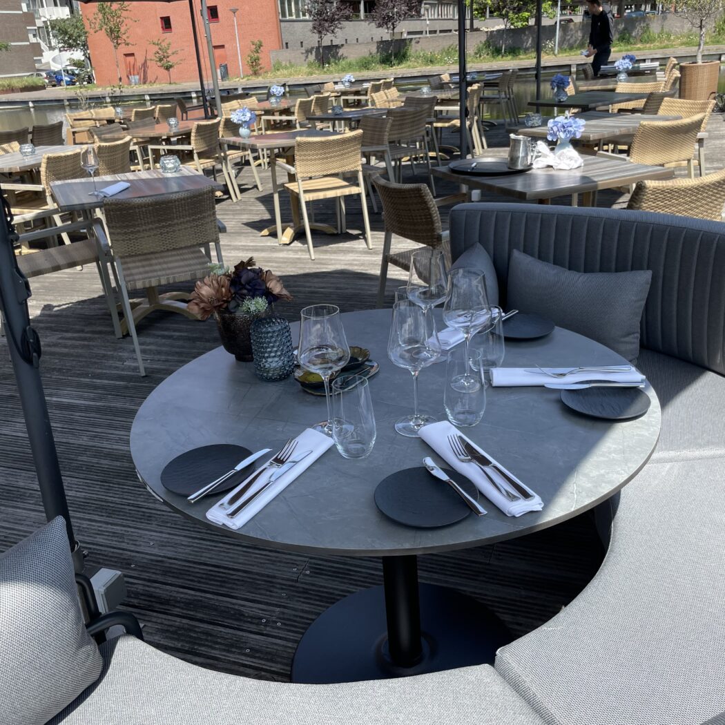 Zonnig restaurantterras met gedekte tafels en comfortabele grijze zitbanken langs het water.