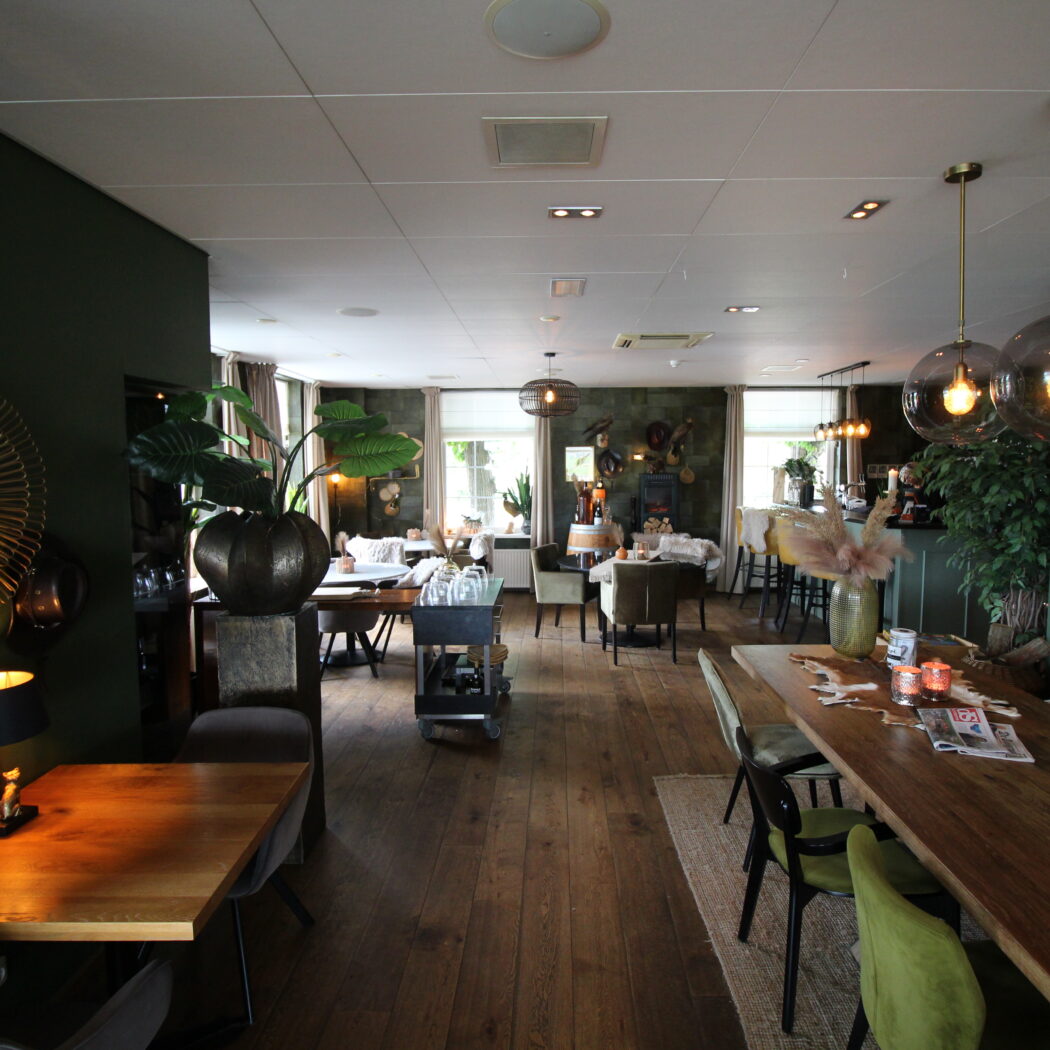 Gezellig restaurant met donkergroene muren, houten vloer, diverse eettafels en comfortabele stoelen onder sfeervolle verlichting.