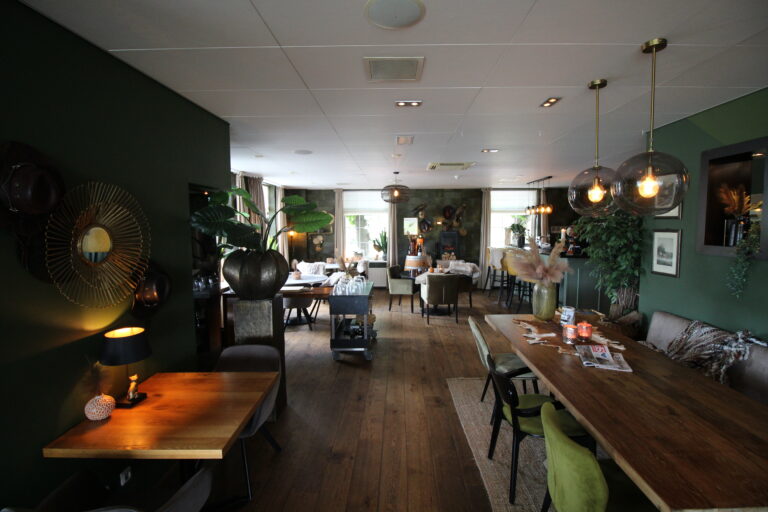 Gezellig restaurant met donkergroene muren, houten vloer, diverse eettafels en comfortabele stoelen onder sfeervolle verlichting.