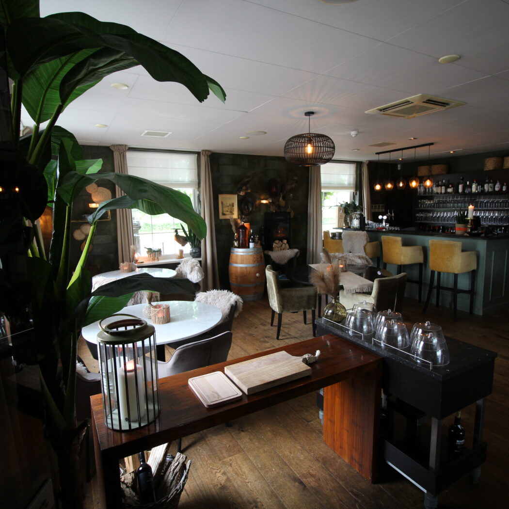 Warm verlicht restaurantinterieur met een bar, gele barkrukken, gezellige tafels en een grote plant.