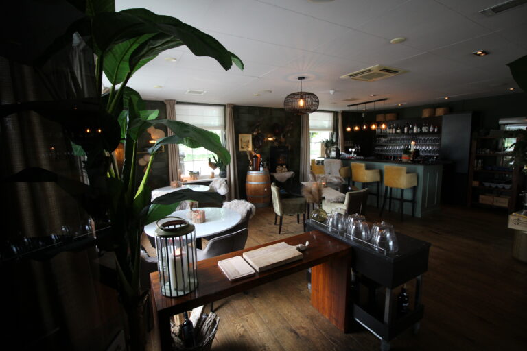 Warm verlicht restaurantinterieur met een bar, gele barkrukken, gezellige tafels en een grote plant.