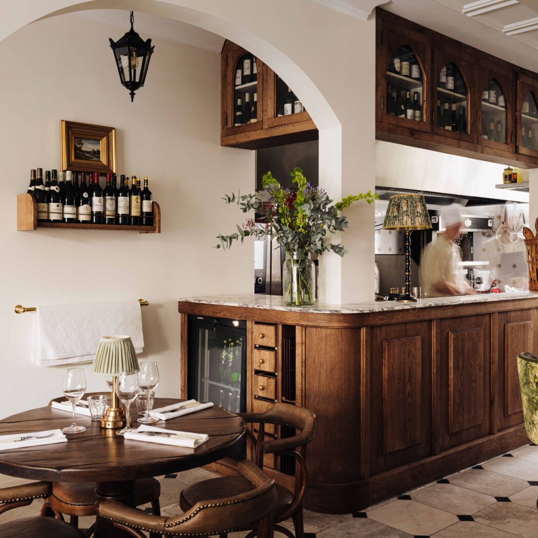 Gezellig restaurant interieur met ronde houten tafel, wijnflessen op planken, een bar en een chef in de open keuken.