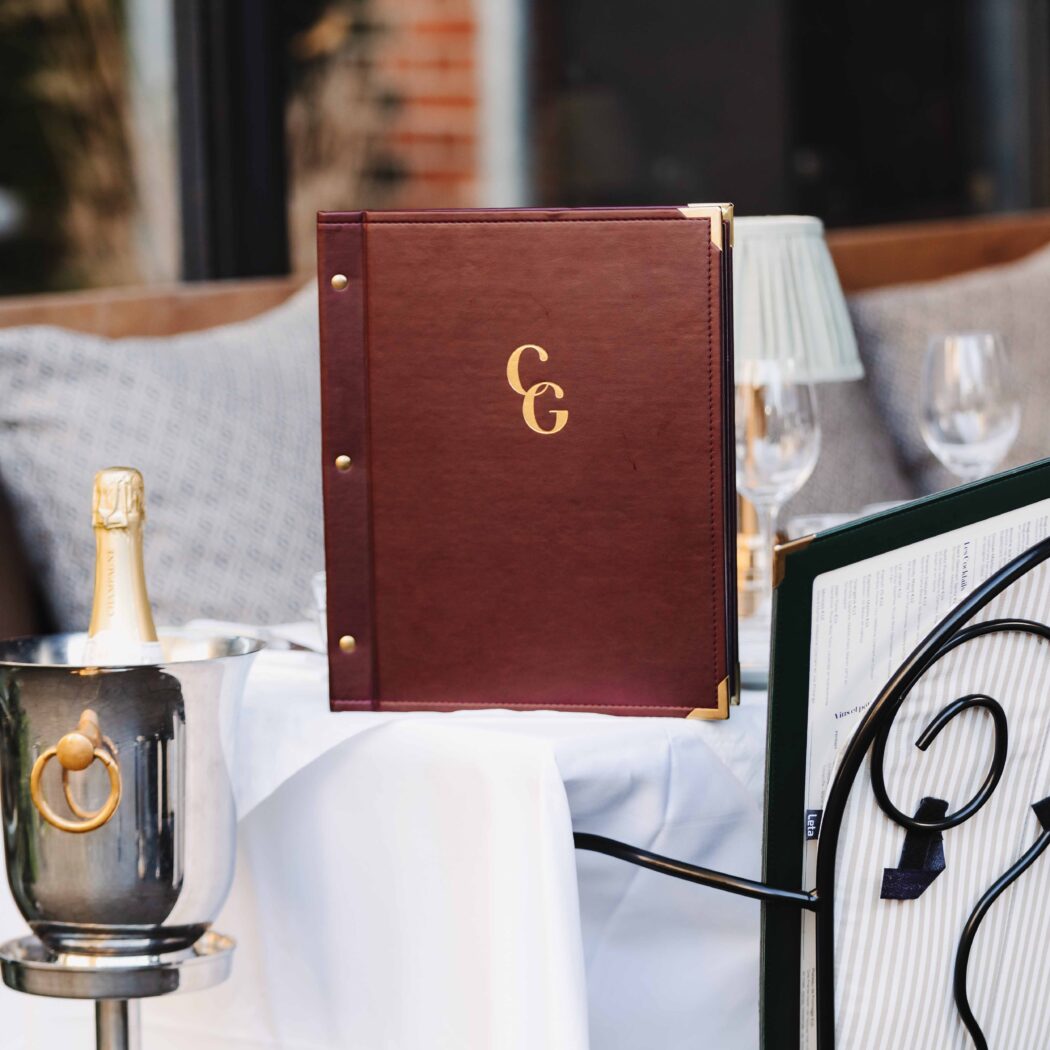 Donkerrood leren restaurantmenu met gouden CG op een witte tafelkleed, naast een champagnekoeler.