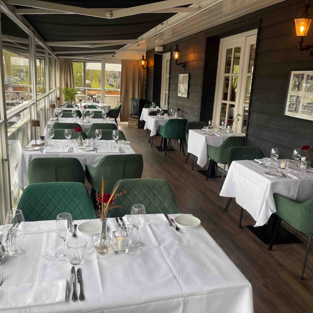 Ruime restaurantzaal met witgedekte tafels, groene fluwelen stoelen en glazen wanden met uitzicht.