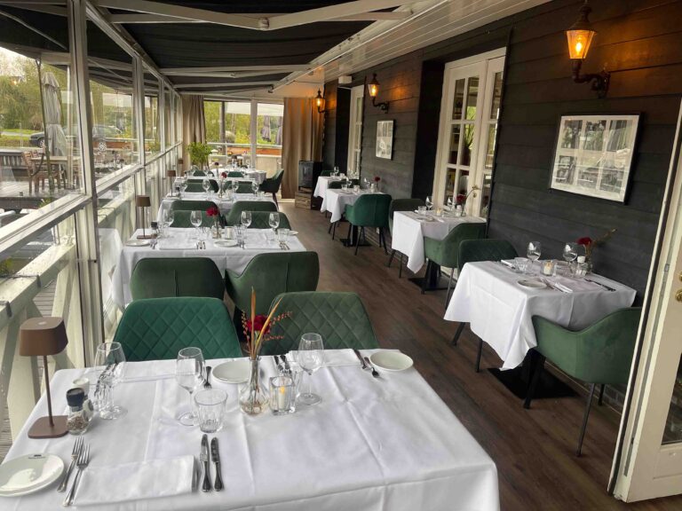 Ruime restaurantzaal met witgedekte tafels, groene fluwelen stoelen en glazen wanden met uitzicht.