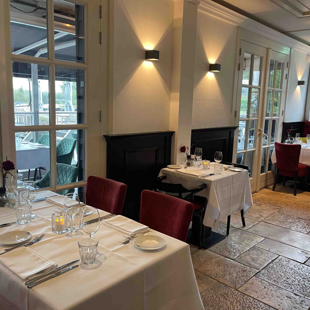 Elegant restaurantinterieur met gedekte tafels, rode fluwelen stoelen en glazen deuren naar een terras.