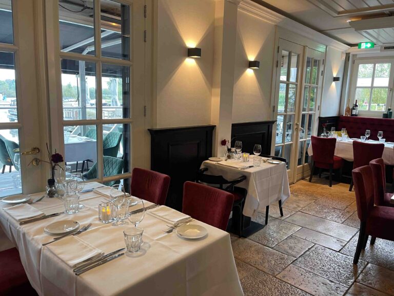 Elegant restaurantinterieur met gedekte tafels, rode fluwelen stoelen en glazen deuren naar een terras.