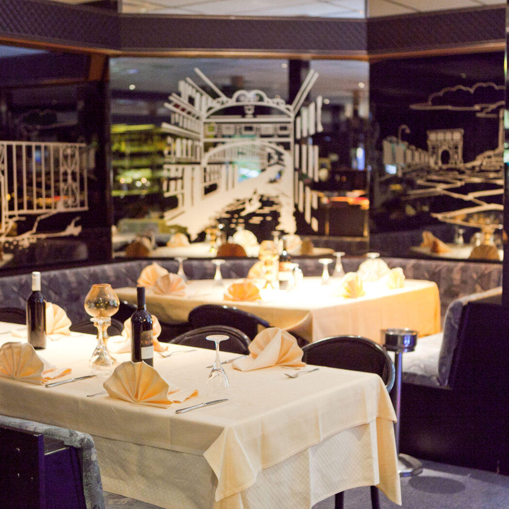 Sfeervol restaurant met gedekte tafels, oranje servetten, wijnflessen en zwarte muren met witte stadsillustraties.