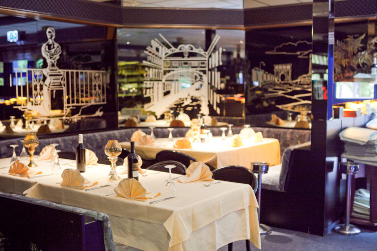 Sfeervol restaurant met gedekte tafels, oranje servetten, wijnflessen en zwarte muren met witte stadsillustraties.