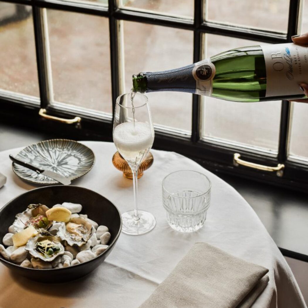 Iemand schenkt Cava OR uit fles in flûteglas, naast verse oesters en bestek op een witte tafel.