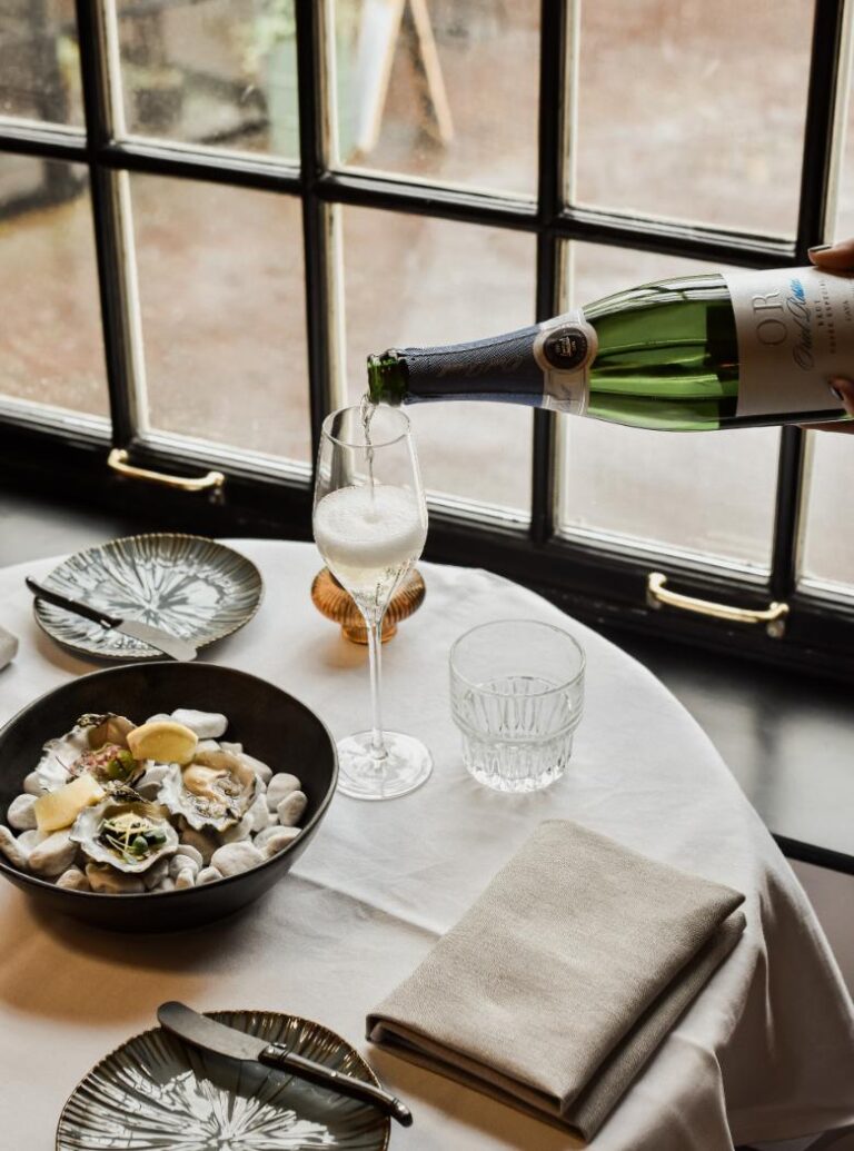 Iemand schenkt Cava OR uit fles in flûteglas, naast verse oesters en bestek op een witte tafel.