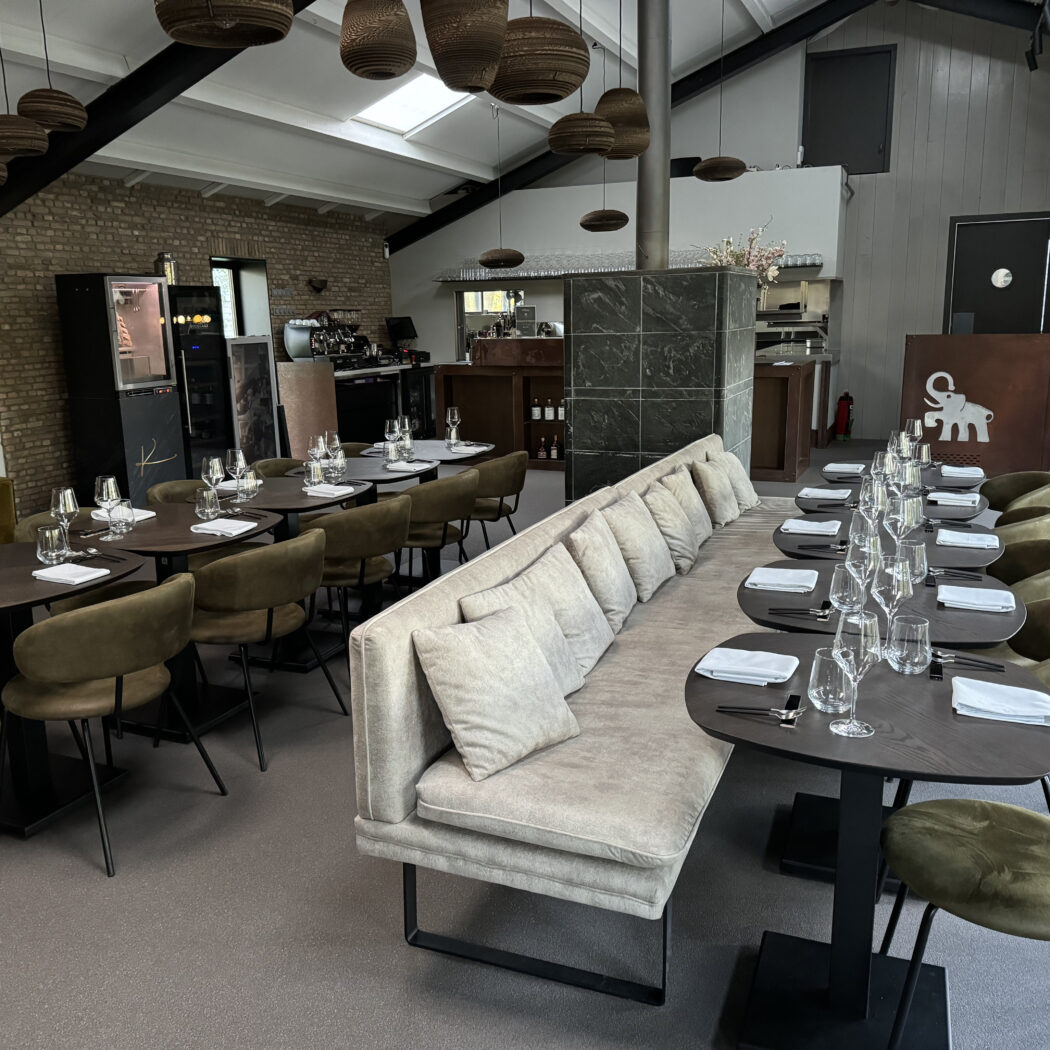 Modern restaurantinterieur met gedekte tafels, olijfgroene stoelen, lange bank en een olifantlogo.