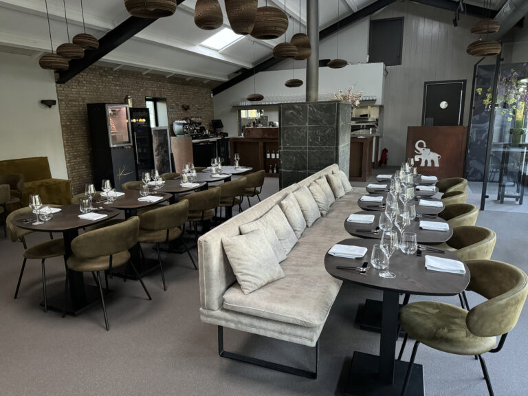 Modern restaurantinterieur met gedekte tafels, olijfgroene stoelen, lange bank en een olifantlogo.