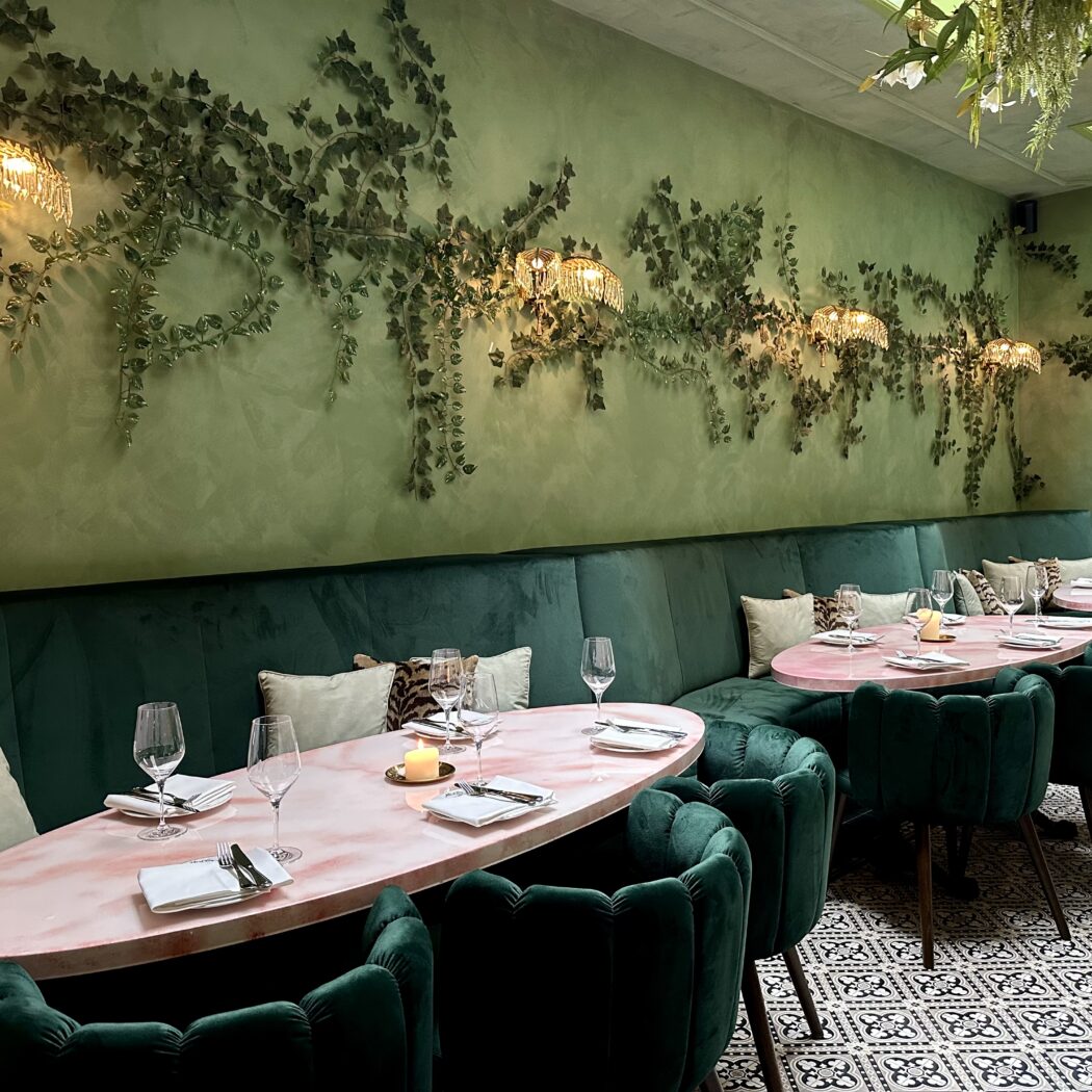 Restaurant interieur: groene fluwelen banken, roze tafels, hangende planten en kristallen wandlampen.