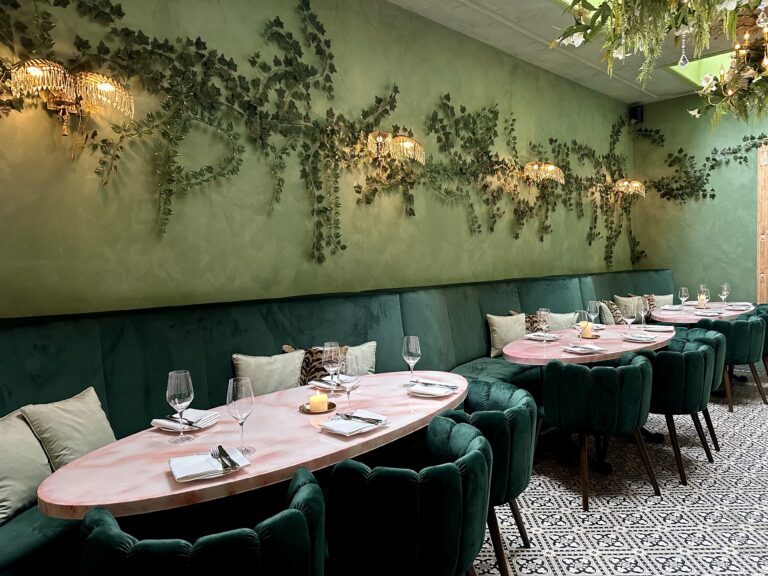 Restaurant interieur: groene fluwelen banken, roze tafels, hangende planten en kristallen wandlampen.