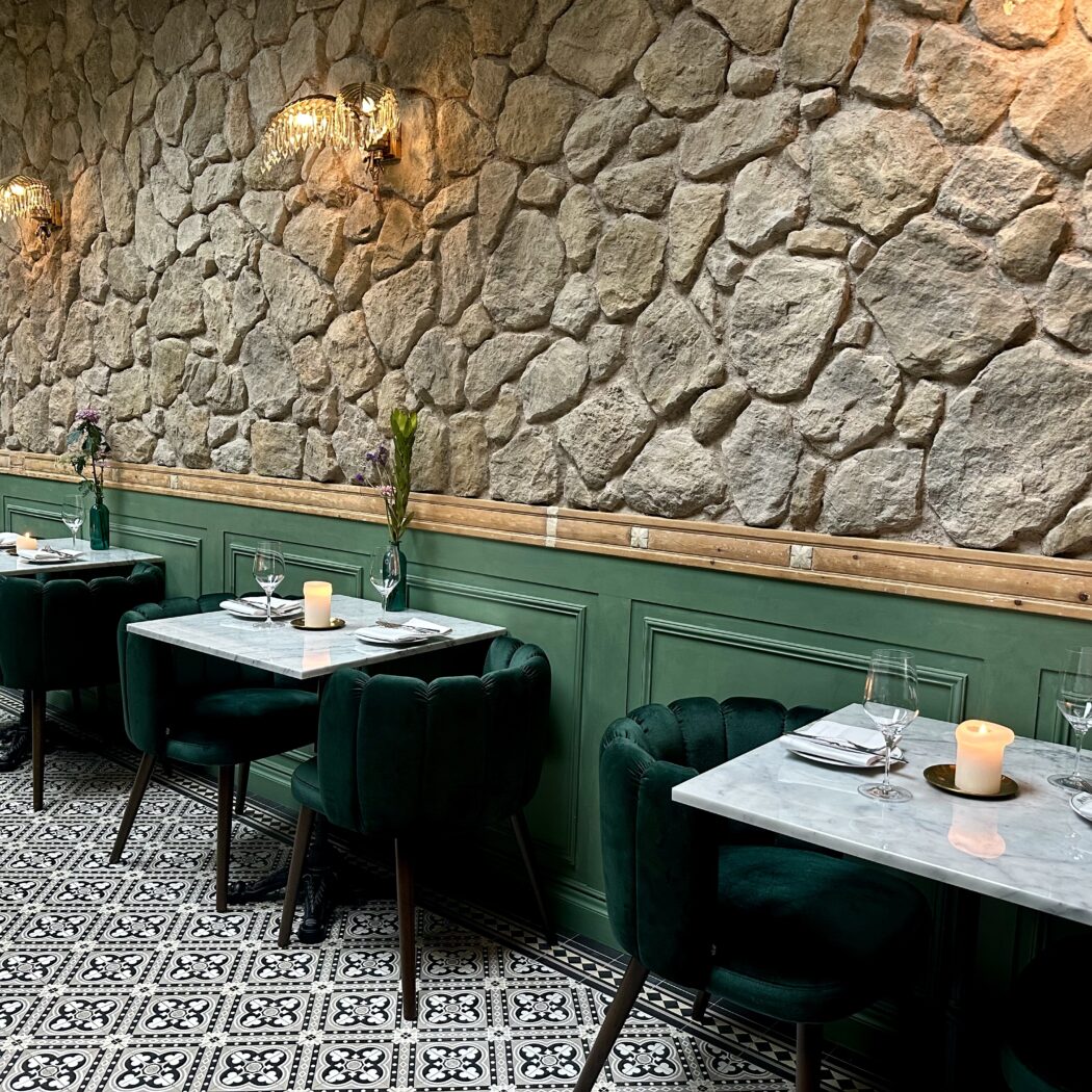 Restaurantinterieur: groene fluwelen stoelen, marmeren tafels, stenen wand en betegelde vloer.