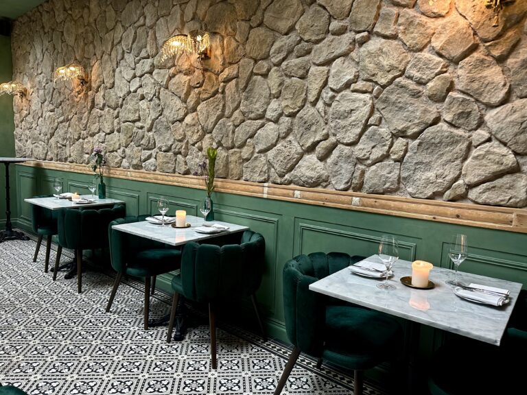 Restaurantinterieur: groene fluwelen stoelen, marmeren tafels, stenen wand en betegelde vloer.