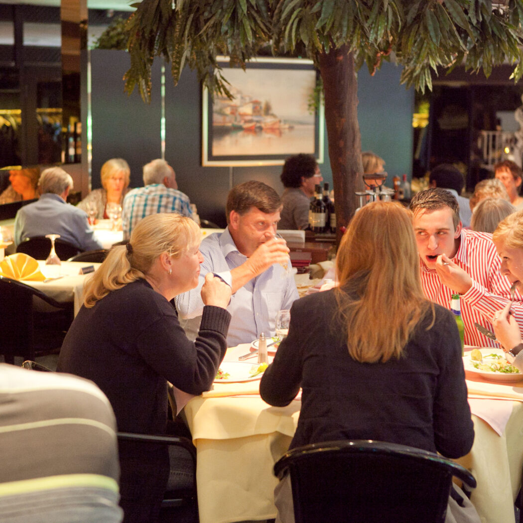 Gasten dineren en praten aan tafels in een restaurant met witte tafellakens en een boomdecoratie.