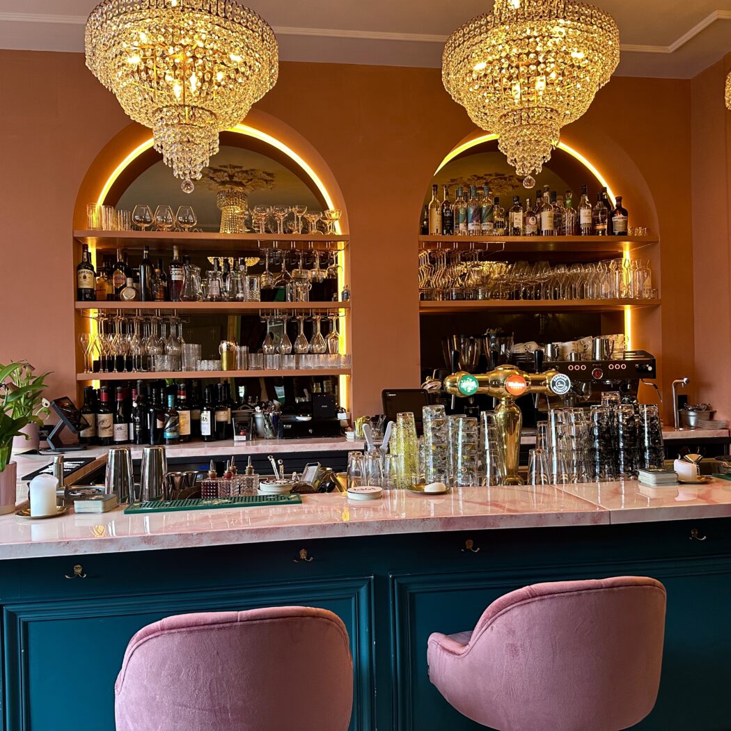 Barinterieur: roze bar met donkere groene kasten, glazen, dranken en kristallen kroonluchters.