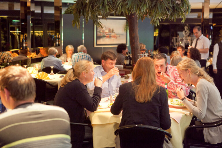 Gasten dineren en praten aan tafels in een restaurant met witte tafellakens en een boomdecoratie.
