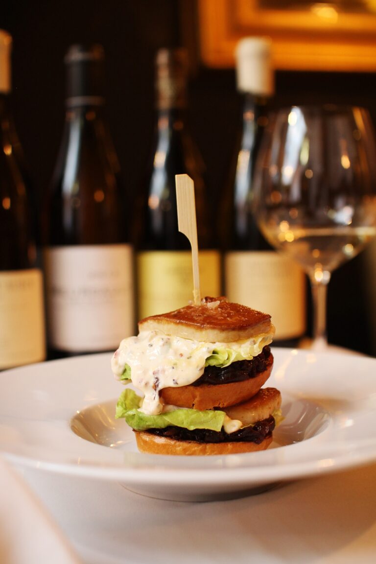 Op een wit bord een luxe mini-burger met gebakken foie gras, sla, jam en romige saus, met flessen wijn erachter.