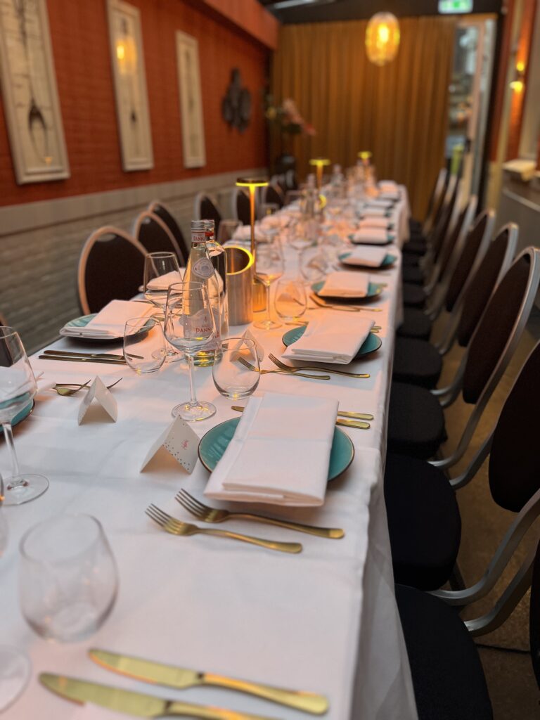 Lange dinertafel feestelijk gedekt met witte lakens, turquoise borden, gouden bestek, waterflessen en sfeervolle verlichting.