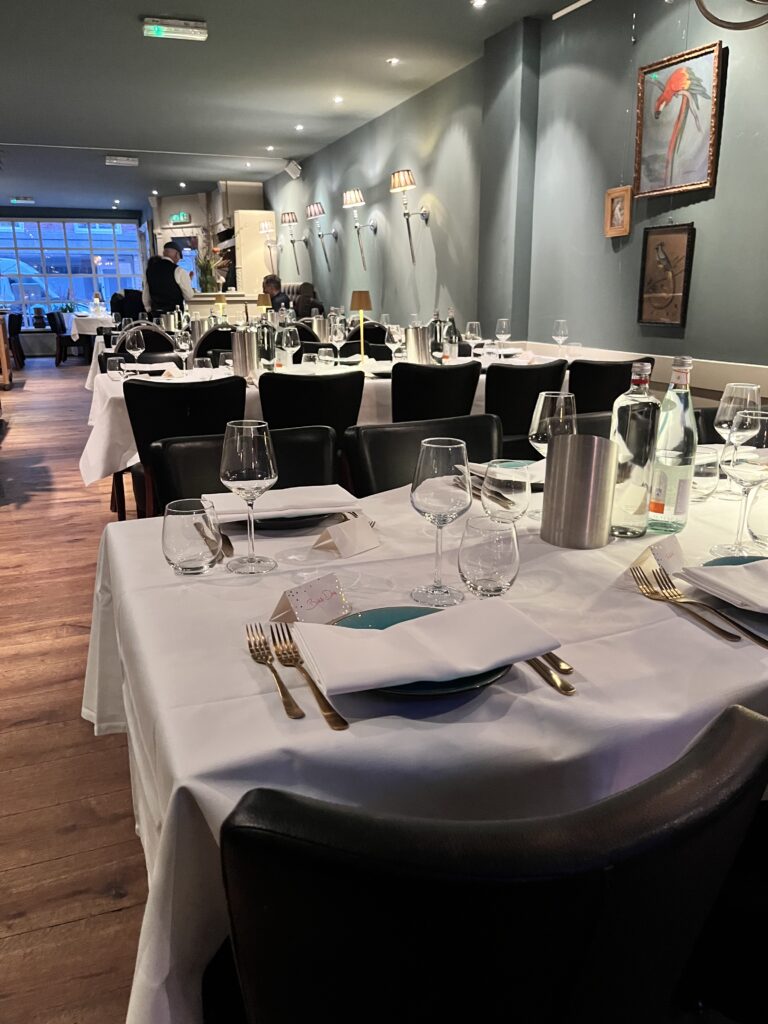 Sfeervol restaurant met opgedekte tafels, gouden bestek, teal borden en wandlampen langs een blauwgrijze muur.