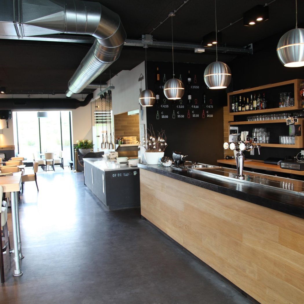 Modern restaurant met houten bar, industriële elementen en een zwarte muur met krijtbordagenda en prijzen.