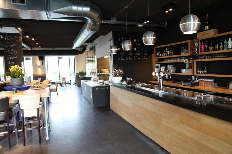 Modern restaurant met houten bar, industriële elementen en een zwarte muur met krijtbordagenda en prijzen.
