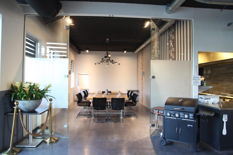 Moderne vergaderruimte met houten tafel en zwarte stoelen, zichtbaar door glas, met plant, gouden afzetpaaltjes en zwarte gasbarbecue.