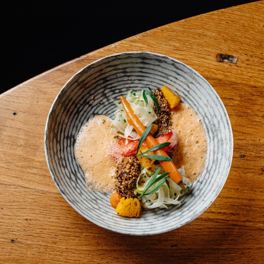 Culinair gerecht met gekookte wortels, witte groenten, rood element, granen en kruiden in gestreepte kom op houten tafel.
