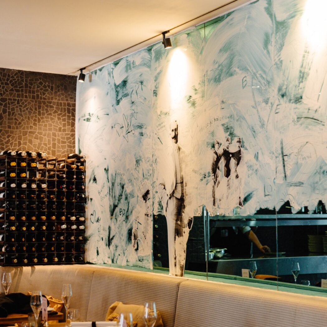 Stijlvol restaurantinterieur met gedekte houten tafels, een lange bank, wijnrekken en een abstracte glazen wand die de keuken weerspiegelt.