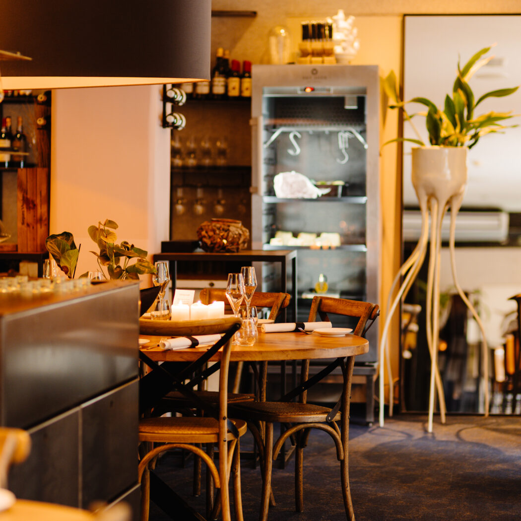 Uitnodigend restaurantinterieur met ronde houten tafel, wijnglazen, brandende kaarsen en een vitrine met lekkernijen.